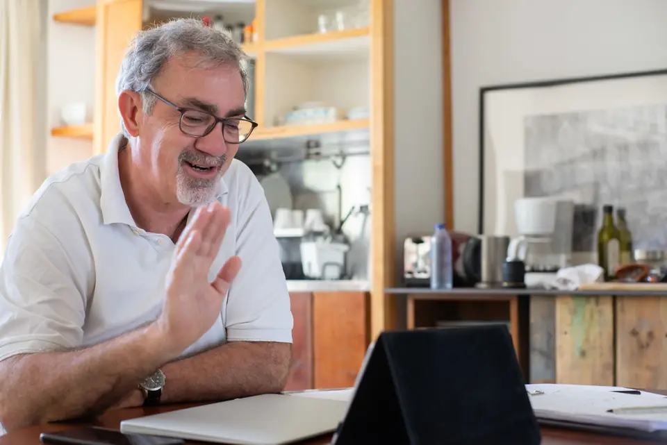 A smiling older man with glasses waves at his tablet during a video call at his kitchen table.