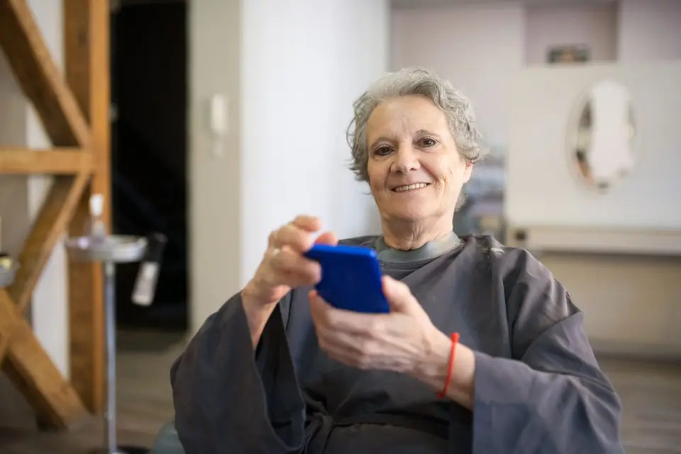An older woman with white hair smiles while holding her smartphone at home.