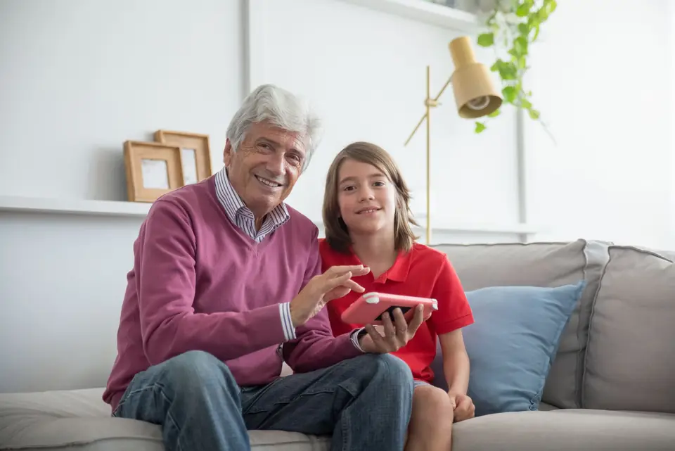 A smiling grandfather and his granddaughter sit together on a sofa, looking at a tablet.