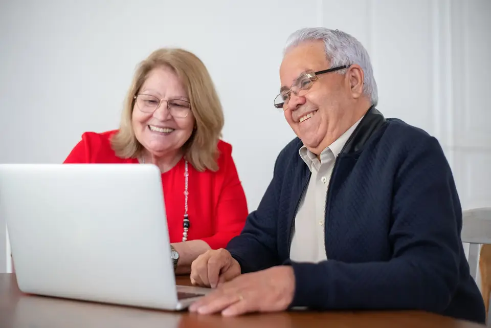 An older couple smile together as they read something on a laptop at their kitchen table.